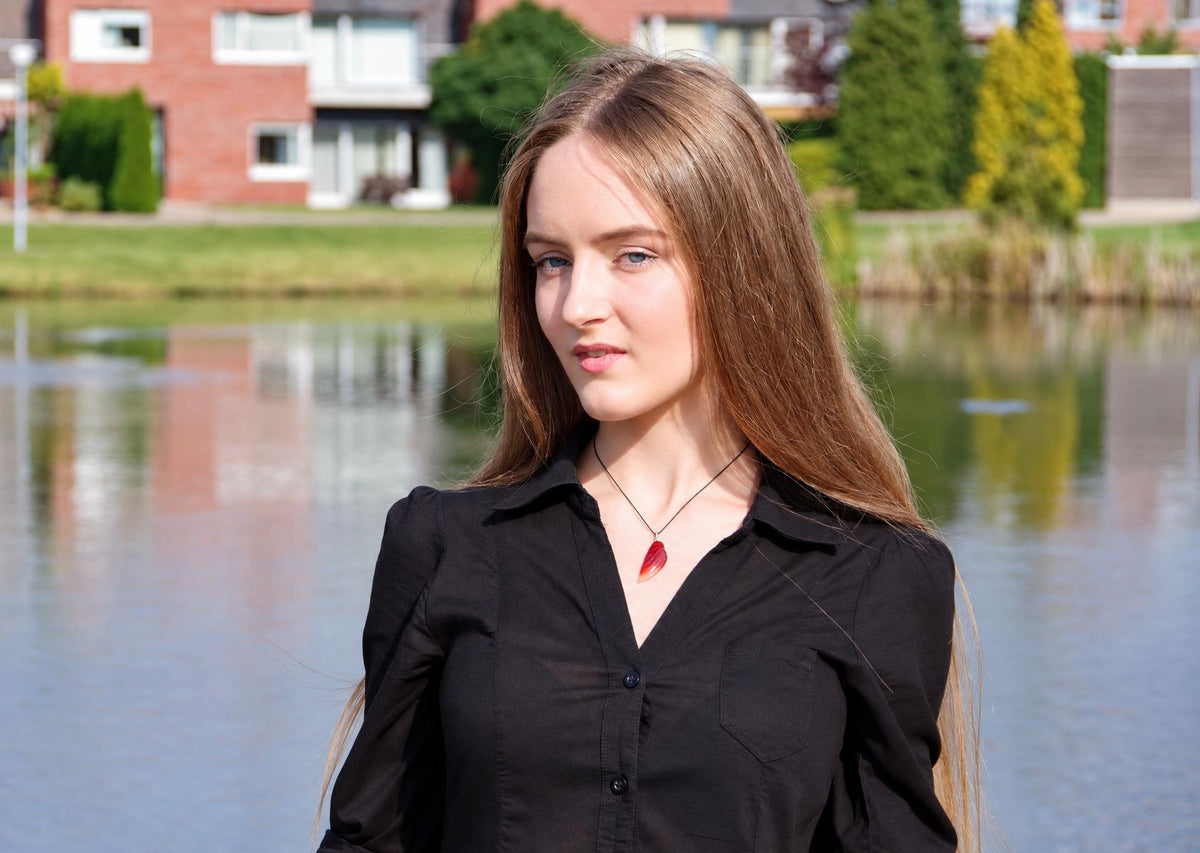 A woman wears a black shirt and a necklace with a red, wing-shaped pendant against a blurred outdoor background.