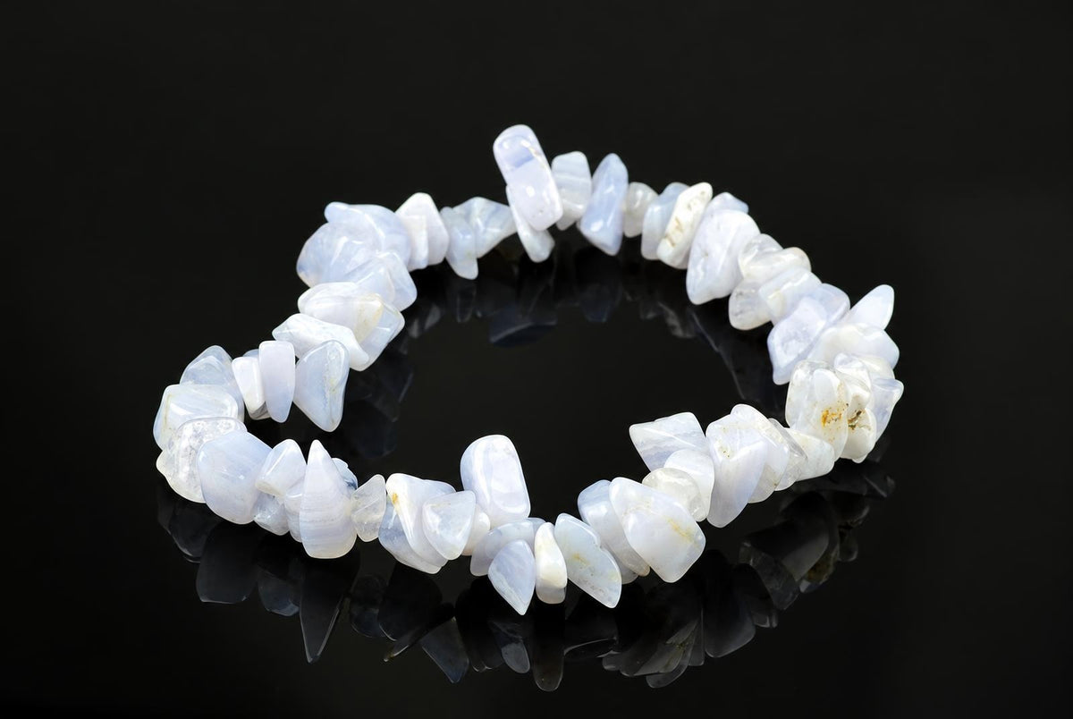 A bracelet made of irregular, polished chalcedony chips in pale blue and white tones rests on a dark reflective surface.