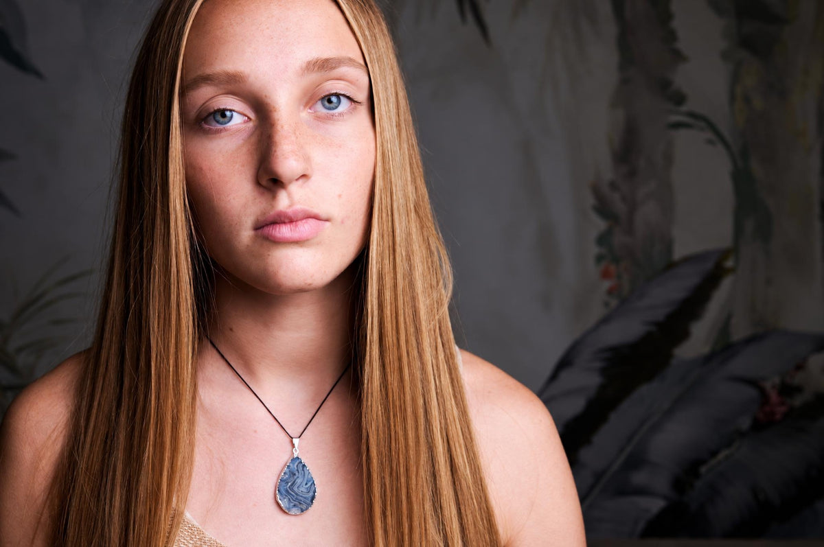 A person wears a teardrop-shaped pendant with blue and white swirls on a dark cord against a dark surface.