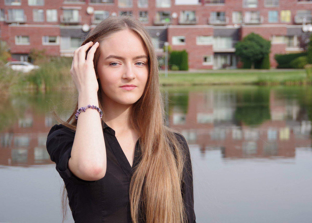 Young woman with long blonde hair wearing a Charoite bracelet – Chips on her wrist, standing outdoors near a calm lake with buildings in the background, natural crystal jewellery.
