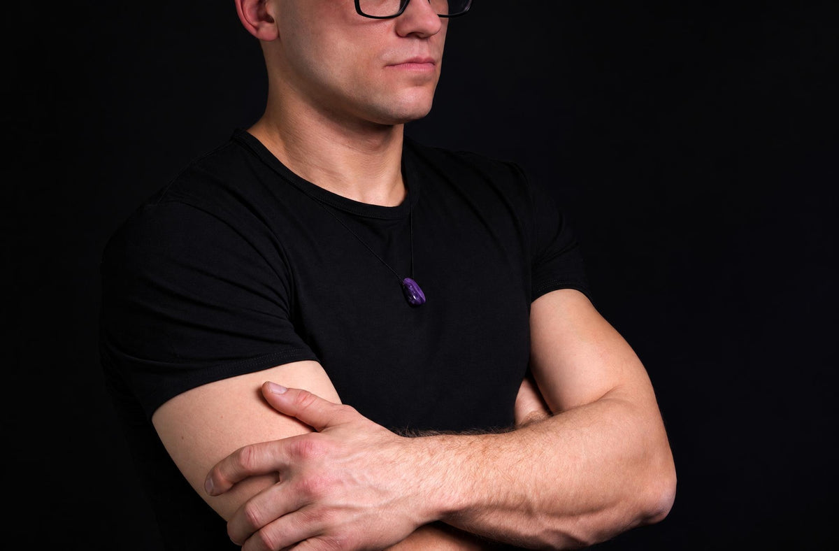 A man in a black t-shirt wears a dark purple drop-shaped pendant on a thin cord against a dark background.