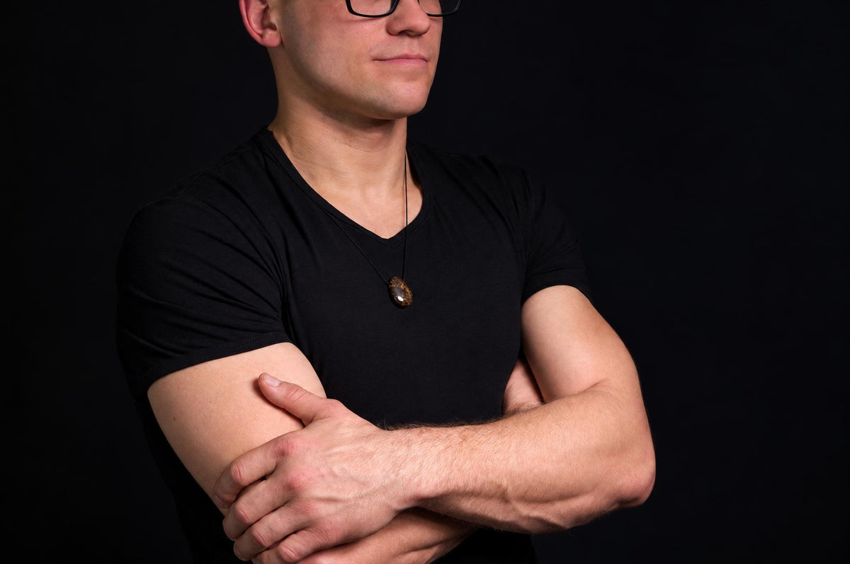 A man in a black t-shirt wears a pendant with a dark brown, polished drop-shaped stone on a thin cord against a dark background.