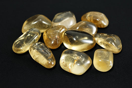 A pile of smooth, polished citrine stones in yellow and clear tones on a dark surface.