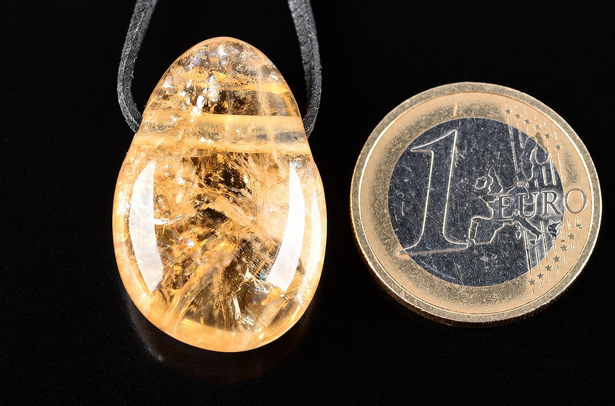 A teardrop-shaped citrine pendant with golden inclusions on a dark cord, next to a one-euro coin on a black surface.