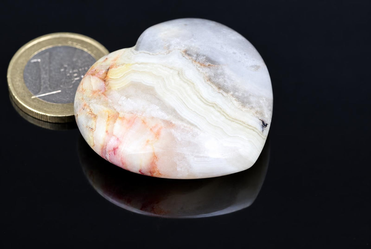 A polished pale heart with wavy cream, grey and peach bands rests beside a coin on a dark reflective surface.