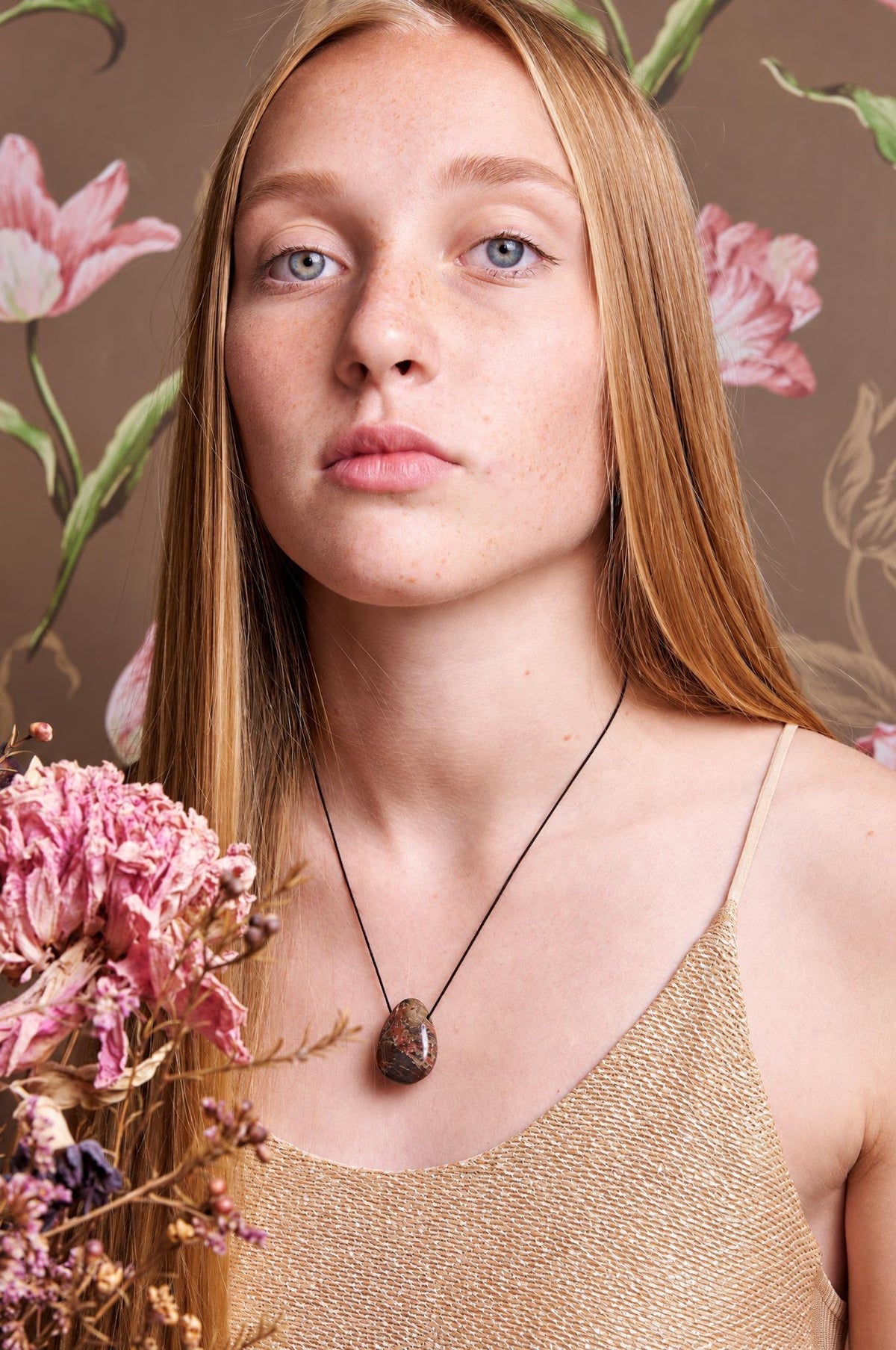 A woman wears a pendant necklace with a dark brown, oval stone on a black cord against a floral background.