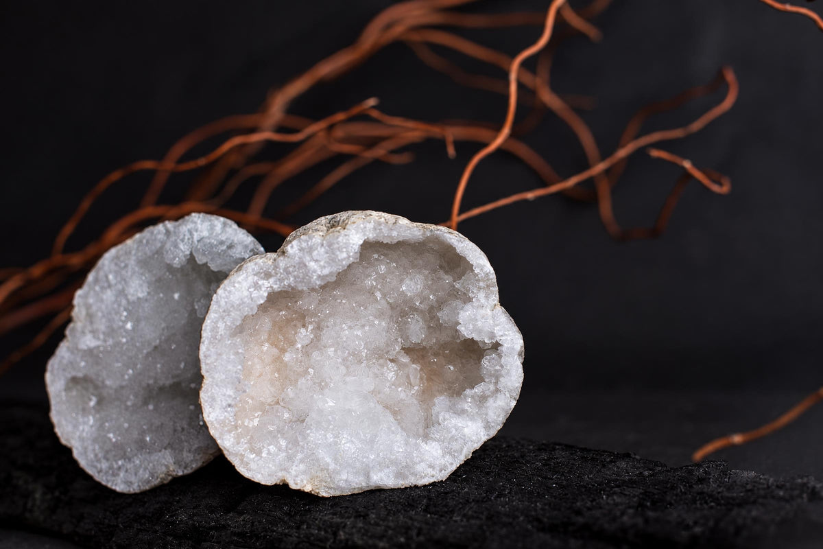 Two white crystal geodes with rough outer shells and sparkling inner crystals rest on a dark surface, near brown twigs.