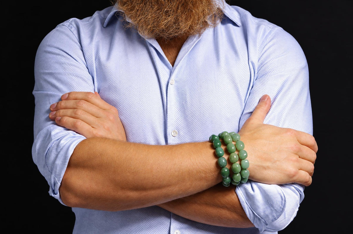 Three rows of polished green beads around a wrist, shown on a person with crossed arms and a pale blue patterned shirt against a dark background.