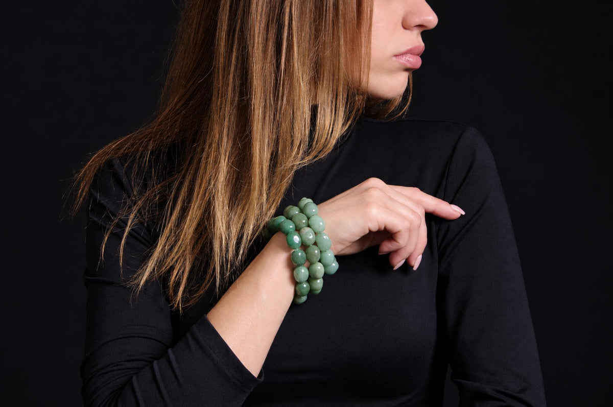 Close-up of glossy rounded green bead bracelets stacked on a model’s raised wrist, with long brown hair and a black top against a dark background.