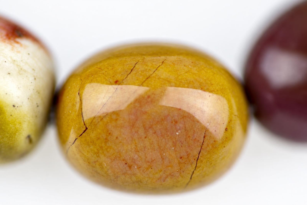 Close-up of a glossy rounded mustard-yellow bracelet bead with peach mottling and fine dark lines, flanked by partially visible cream and burgundy beads on a white background.