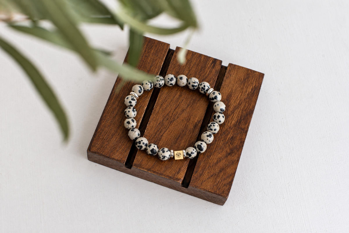 A bracelet with white and black spotted beads, a gold-coloured square charm, and silver-toned spacers rests on a dark wooden surface.