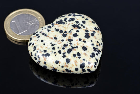 A heart-shaped stone with a cream background and black spots sits on a dark surface next to a coin.