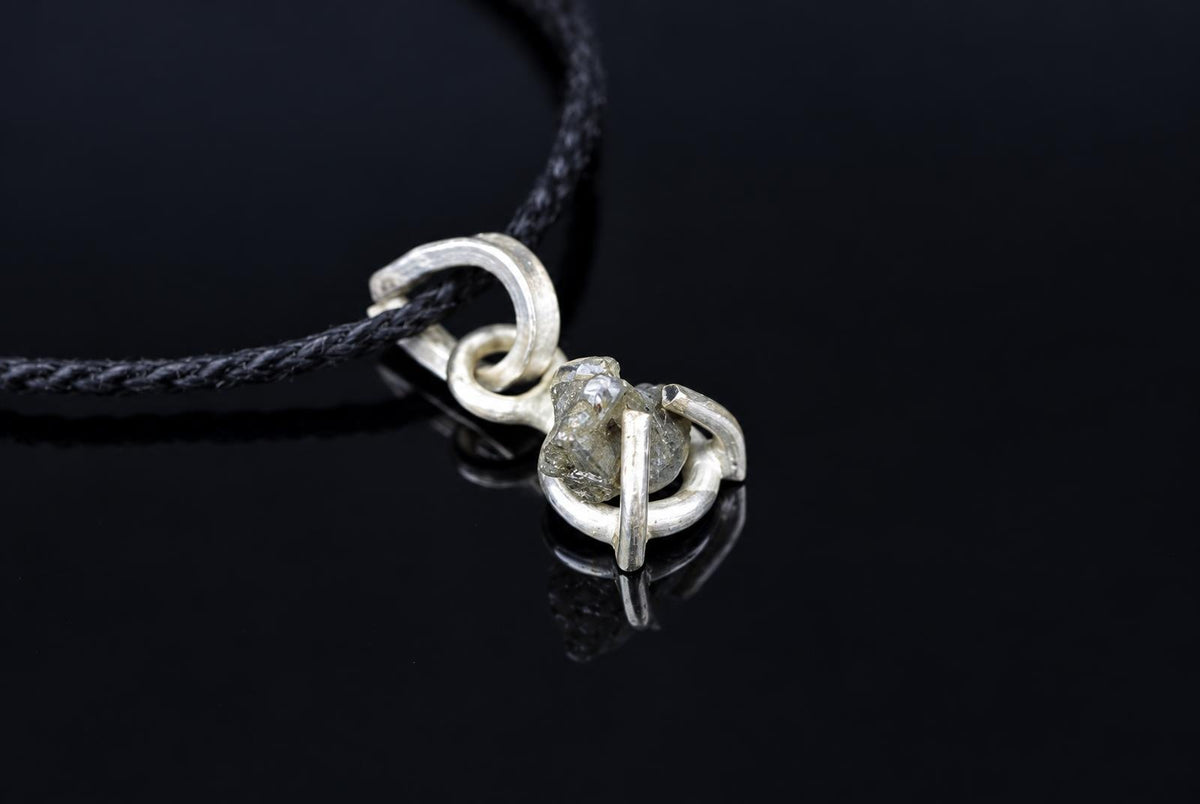 A silver-toned pendant with a rough, greyish stone set in an open loop design on a black cord against a dark surface.