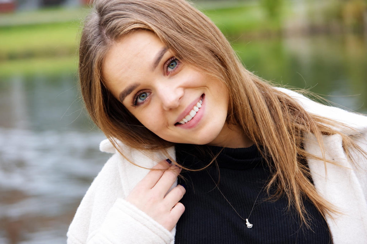 A smiling woman with long brown hair wears a black top and white coat, accessorized with a silver-toned pendant necklace against a blurred outdoor background.