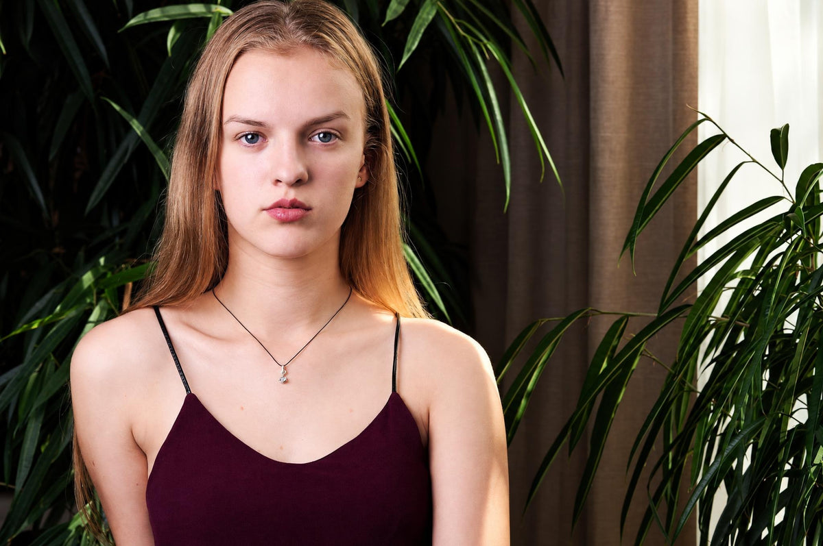 A woman wears a dark purple top and a silver-toned pendant necklace with a small charm, set against green plants.