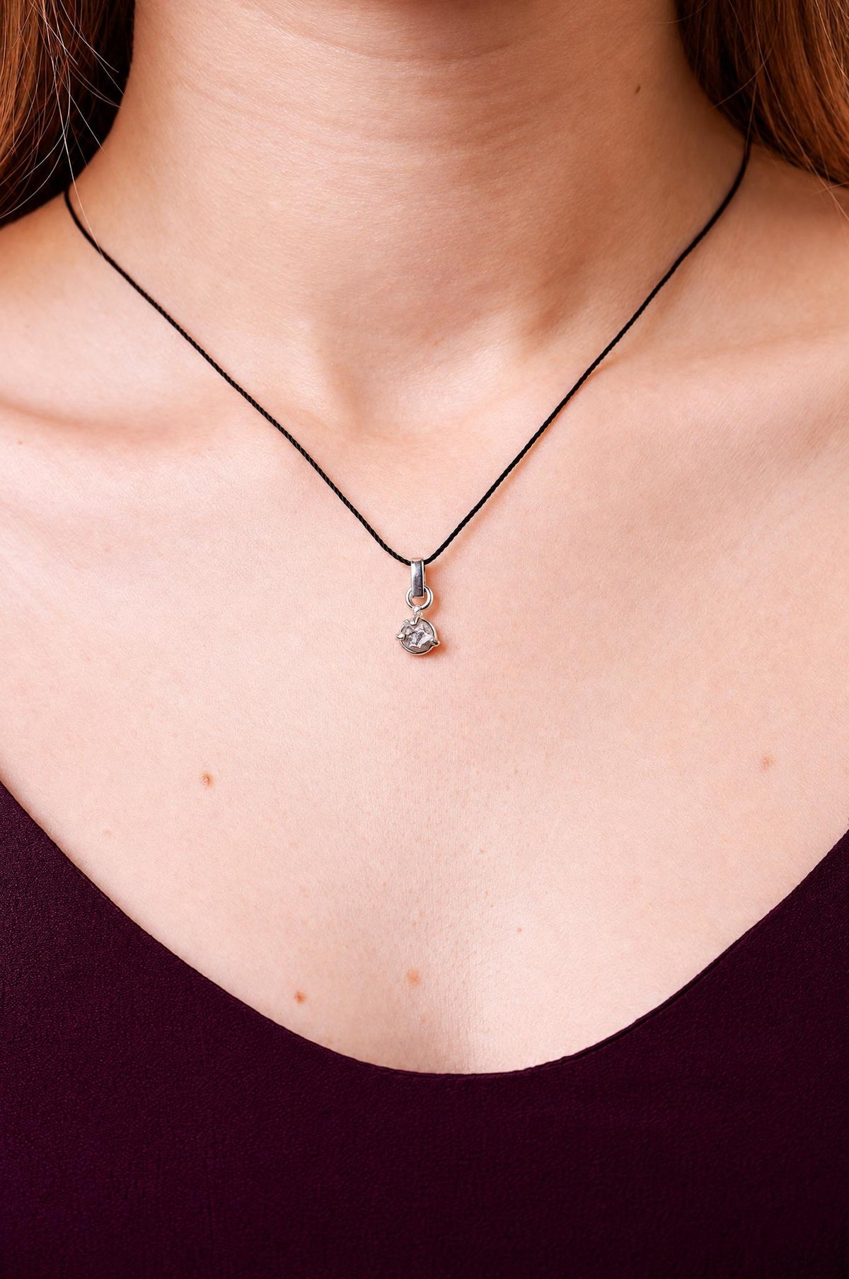 A silver-toned pendant hangs from a black cord on a person's neck, set against a dark top.