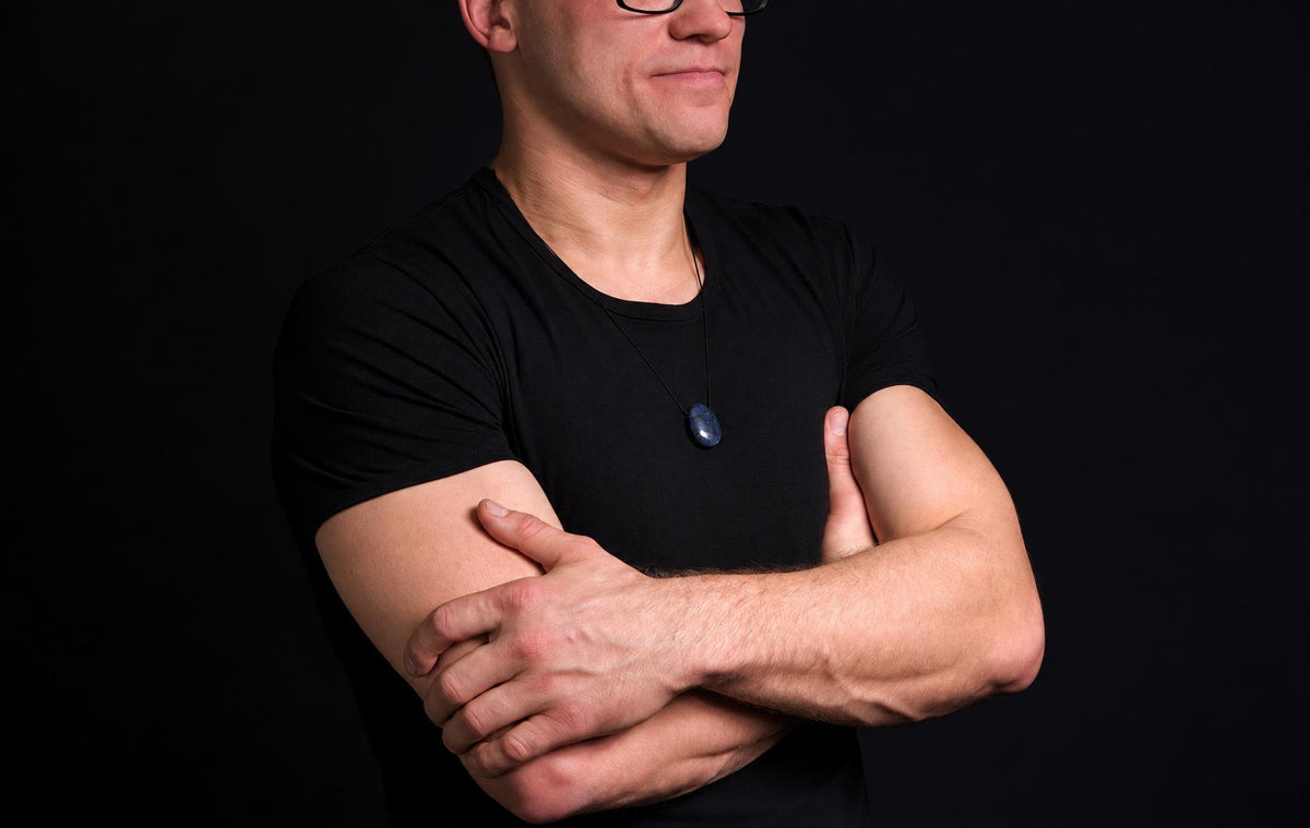 A man in a black t-shirt wears a dark blue oval pendant on a thin cord against a dark background.