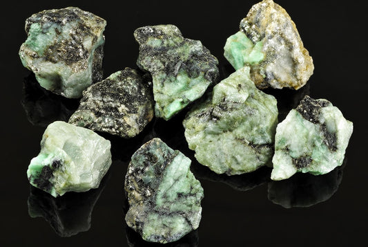 Seven raw emerald specimens with green and black inclusions rest on a dark, reflective surface.
