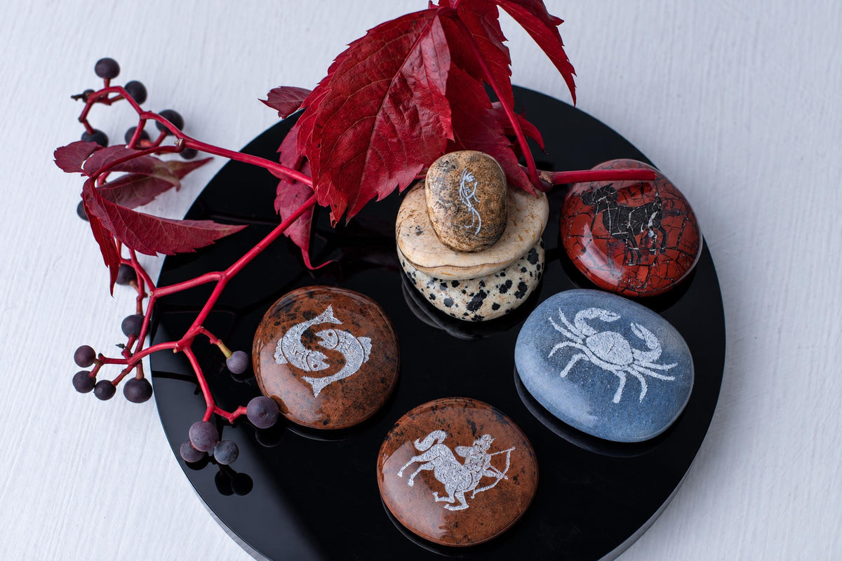 A black circular tray holds several smooth stones with white engravings of zodiac symbols, arranged on a pale wooden surface.