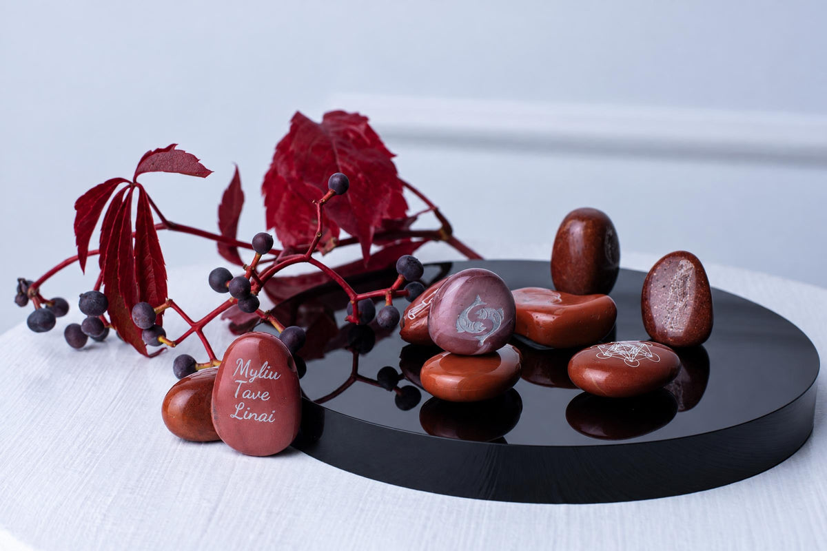 Red polished stones with white engravings rest on a black circular tray, accompanied by red leaves and berries on a pale surface.
