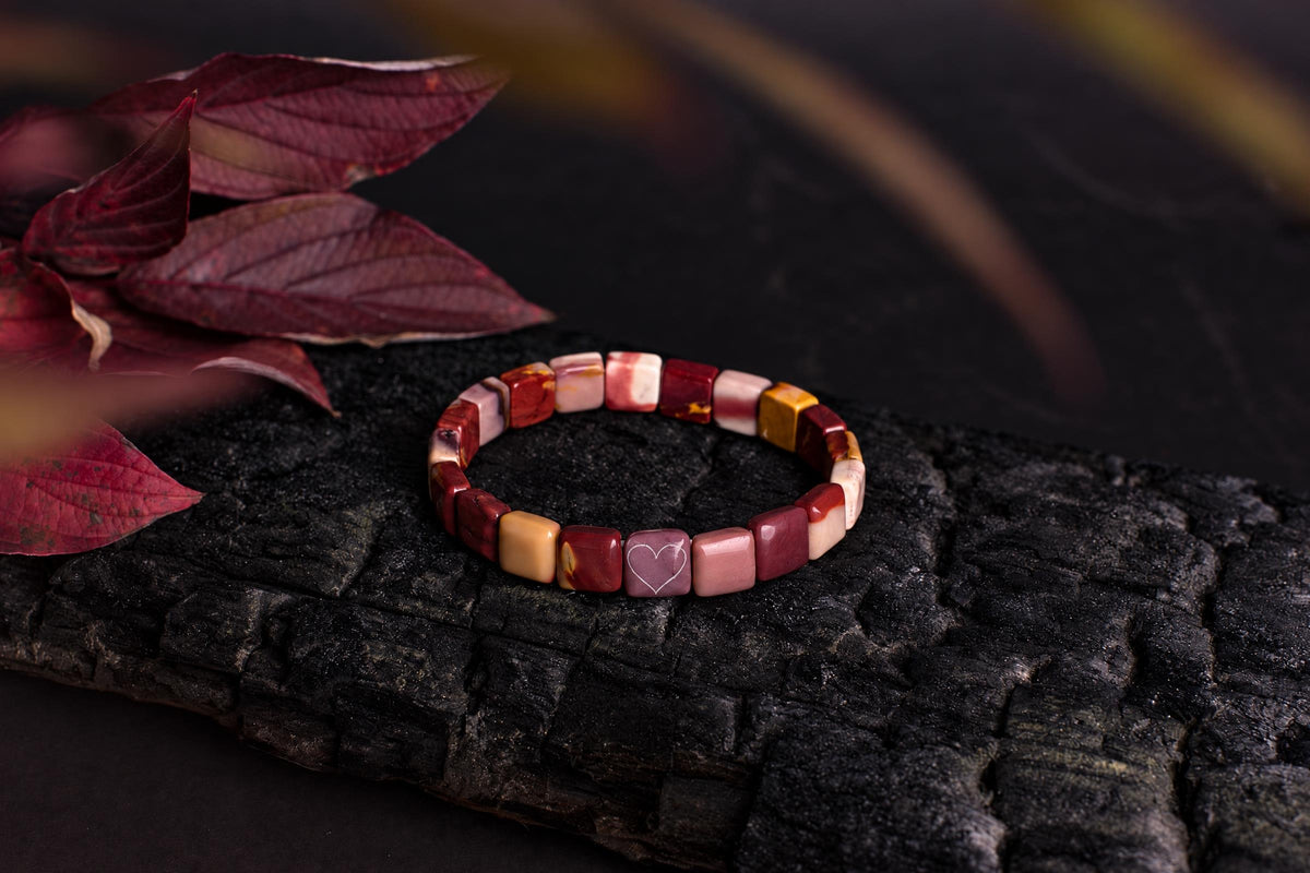 A beaded bracelet with red, pink, and yellow square beads rests on a dark wooden surface next to deep red leaves.