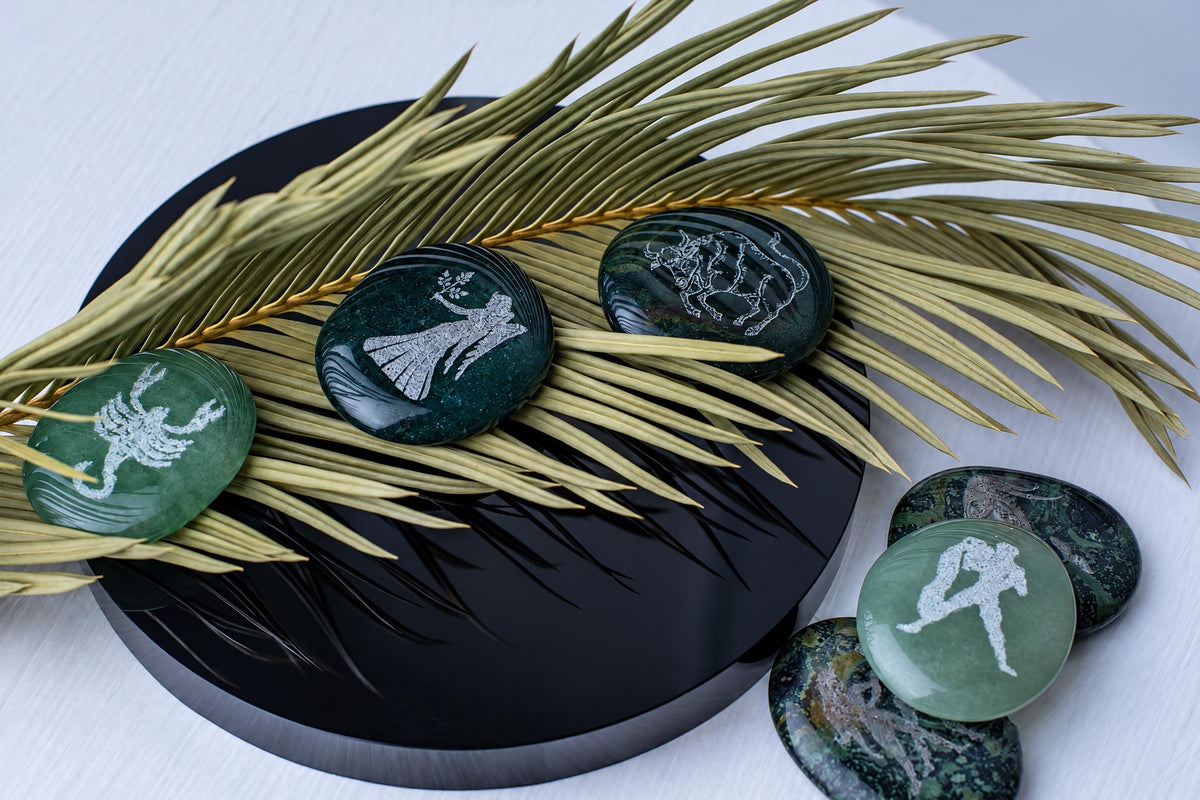 Five polished green stones with white engraved zodiac symbols rest on a dried palm frond atop a dark circular surface.