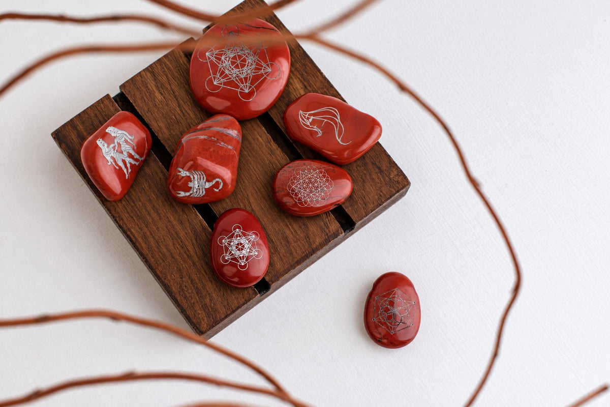 Seven red stones with silver-toned engravings rest on a dark wooden surface and white background.