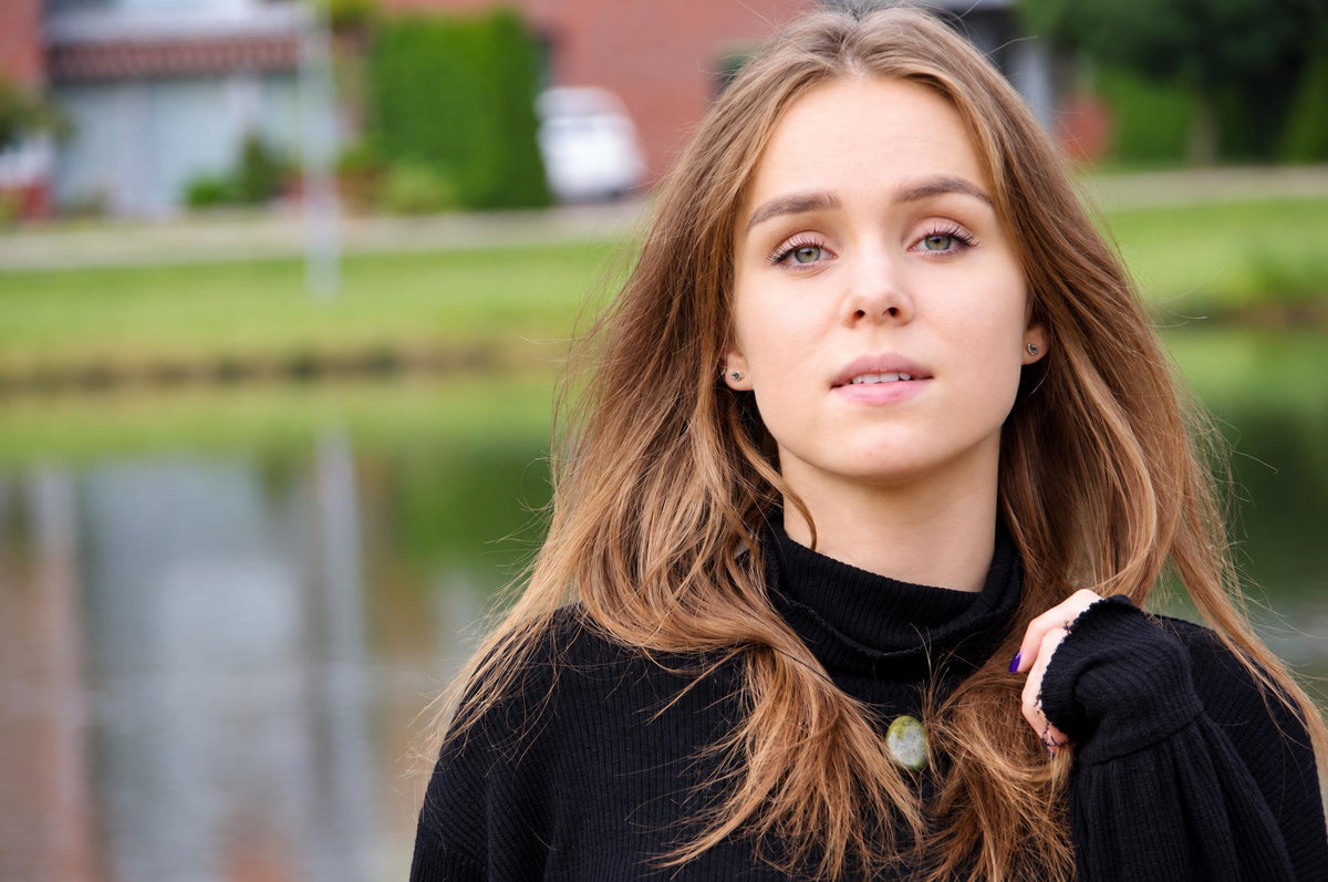 A woman with long brown hair wears a black turtleneck sweater featuring a round green pendant at the neckline.