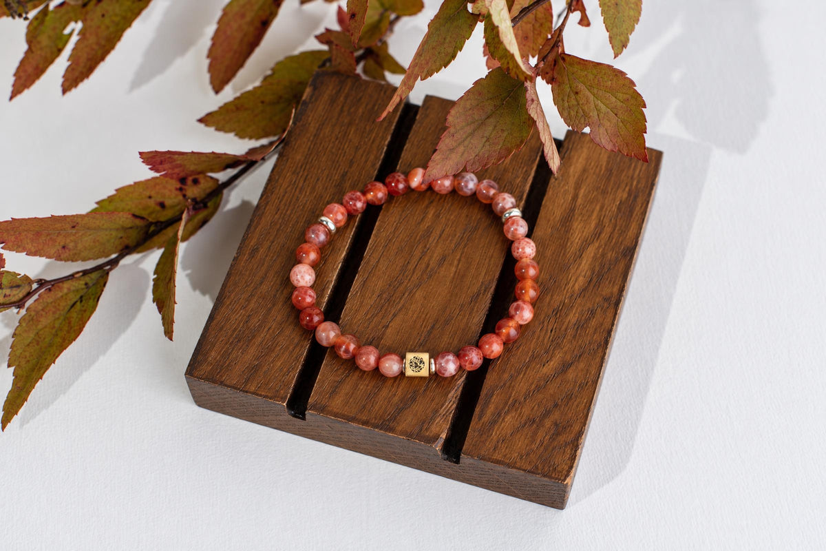 A bracelet with round reddish-orange speckled beads and a gold-toned square charm rests on a dark wooden surface.