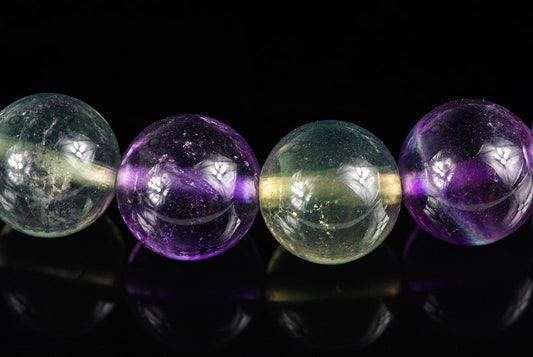 Four round fluorite beads in green and purple hues, arranged horizontally on a dark surface.
