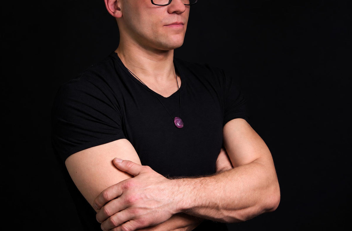 A man in a black t-shirt wears a necklace with a polished, oval-shaped purple stone pendant against a dark background.