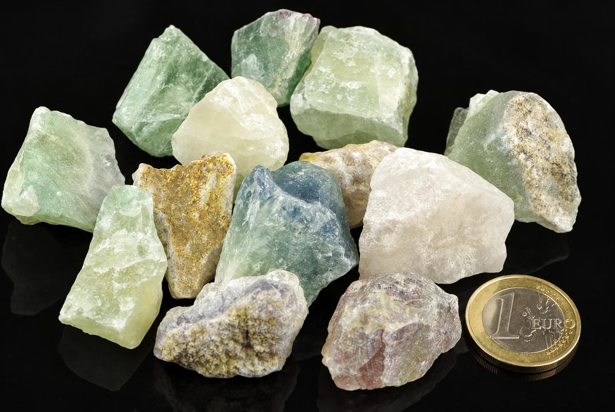A collection of raw fluorite crystals in green, blue, white and yellow tones on a dark surface with a one euro coin for scale.