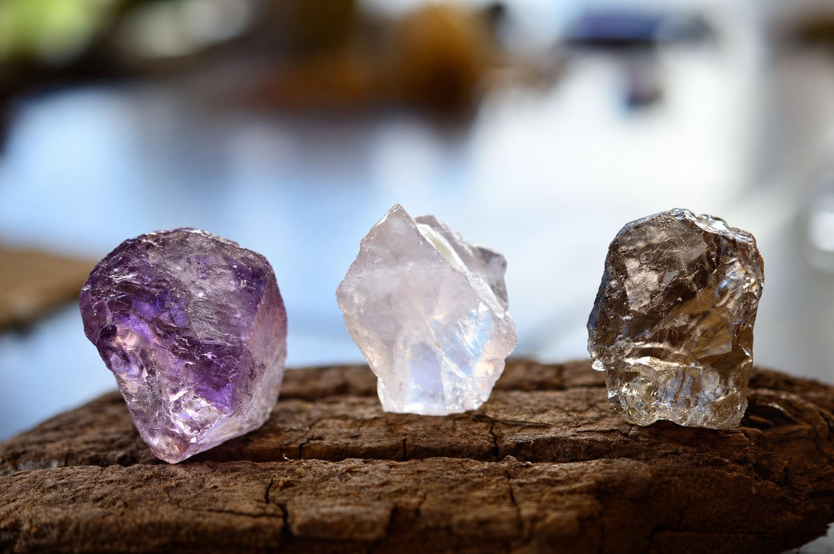Three rough crystals, purple, white and clear, stand on a dark wooden surface against a blurred background.