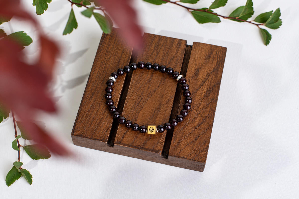 A dark beaded bracelet with a gold-toned square charm and silver spacers rests on a wooden display stand.