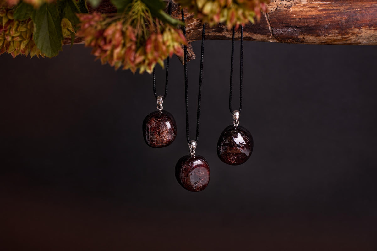 Three round, dark red pendants with silver-toned bails hang from black cords against a dark background.