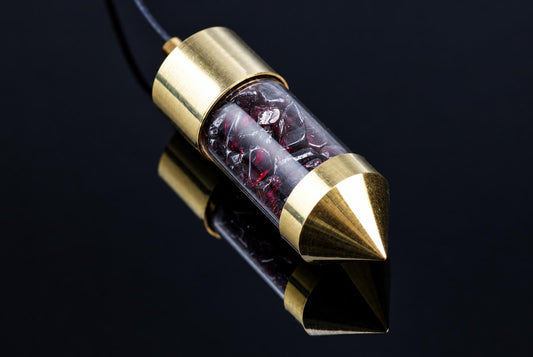 A brass pendulum with a clear tube containing red garnet crystals, resting on a dark reflective surface.