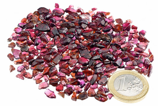 A pile of small, rough-cut garnet stones in shades of red and pink on a white surface next to a one euro coin.