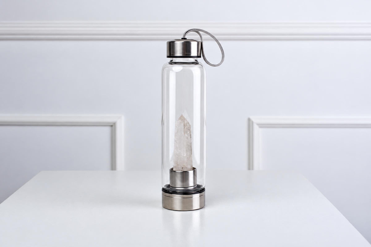 A clear glass water bottle with a silver-toned lid and base, containing a tall, pale quartz crystal inside, standing on a white surface.
