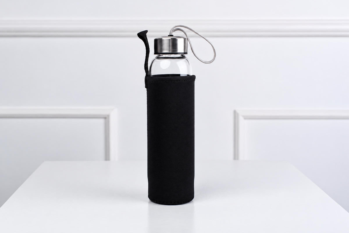 A glass water bottle with a black sleeve and silver-toned lid sits on a white surface.