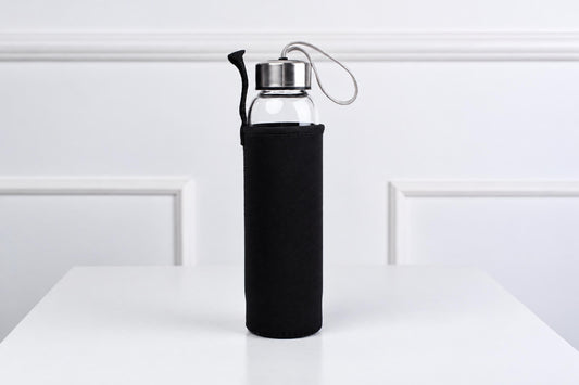 A glass water bottle with a black sleeve and silver-toned lid sits on a white surface.