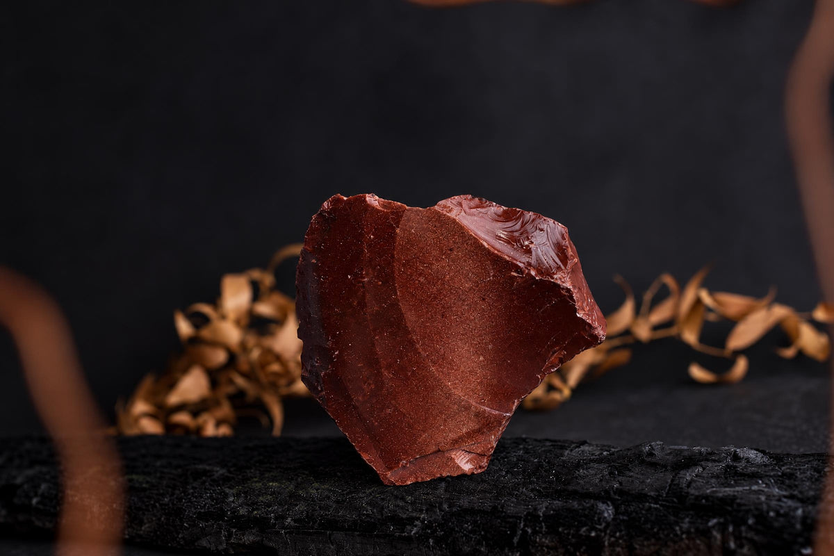 A reddish-brown, irregularly shaped chunk of goldstone rests on a dark surface with dried leaves in the background.