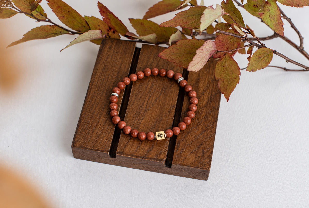 A beaded bracelet with reddish-brown round beads, silver-toned spacers, and a square gold-coloured charm rests on a dark wooden surface.