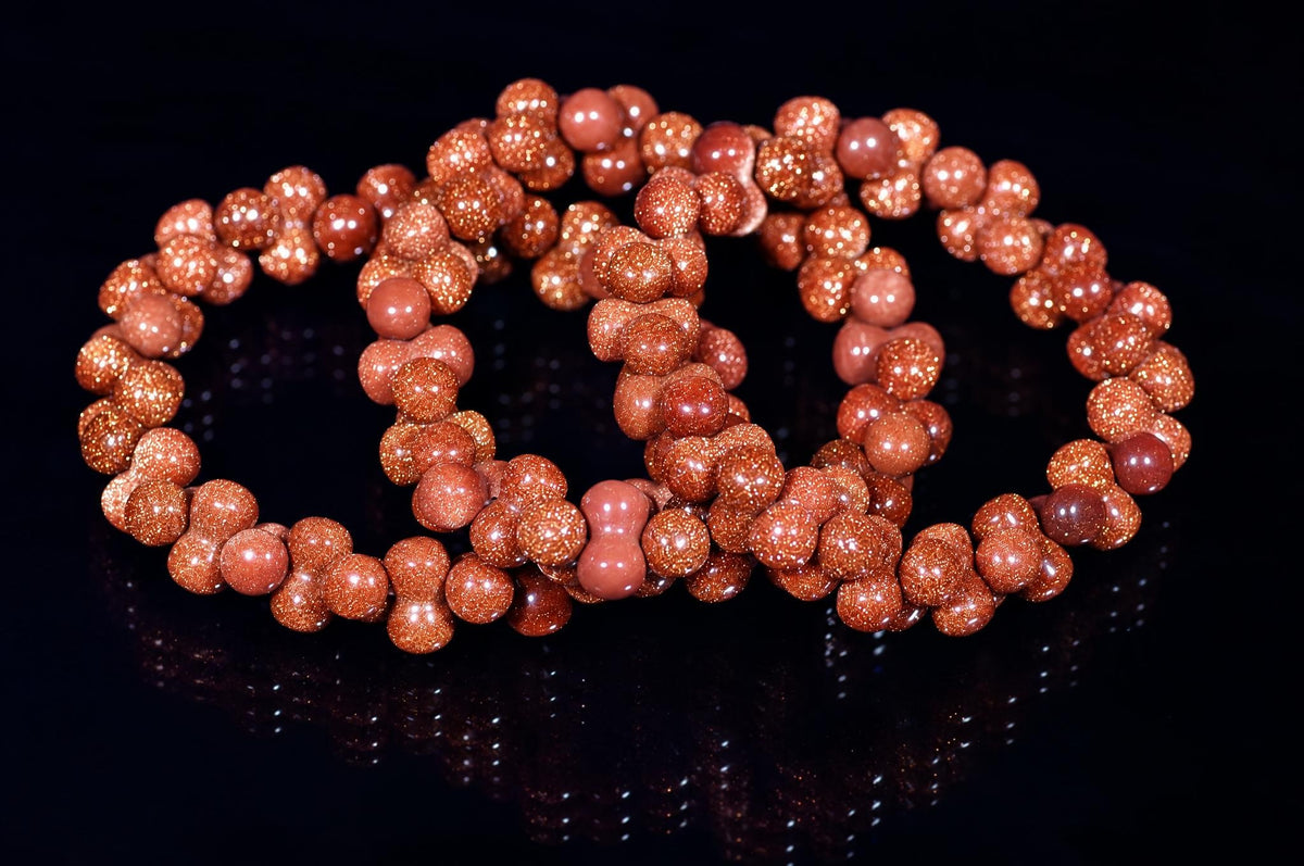 A bracelet made of round, reddish-brown beads with a glittery goldstone finish, arranged in loops on a dark reflective surface.