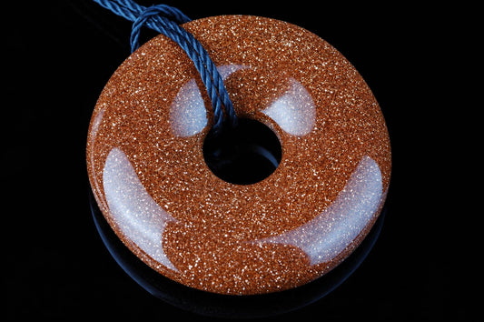 A goldstone pendant with a donut shape, featuring a central hole and white inclusions on a sparkly brown surface, suspended by a blue cord against a dark background.