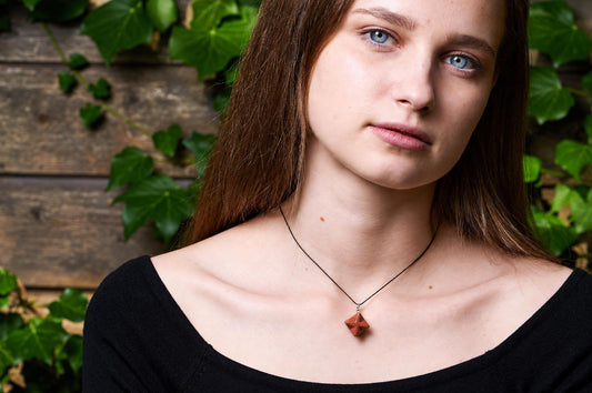 A woman wears a necklace with a reddish-brown, faceted pendant on a black cord against a wooden background with green leaves.