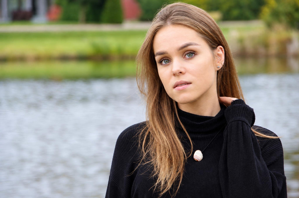A woman wears a black turtleneck sweater with a small, round pendant on a thin cord against a blurred water background.