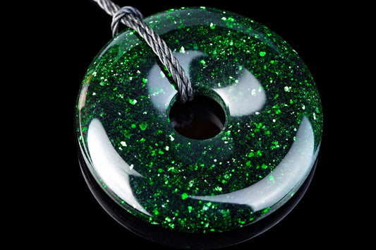 A round, donut-shaped pendant with a glossy green surface speckled with glittering flecks, threaded on a twisted cord against a dark background.