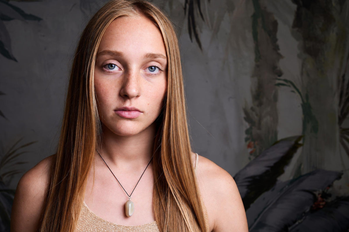 A woman wears a necklace with an oval, pale grey agate pendant on a dark cord against a muted background.