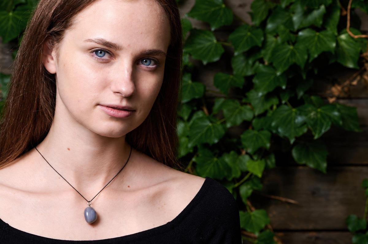 A woman wears a black top and a necklace with a smooth, grey pebble-shaped pendant on a thin cord against green ivy.
