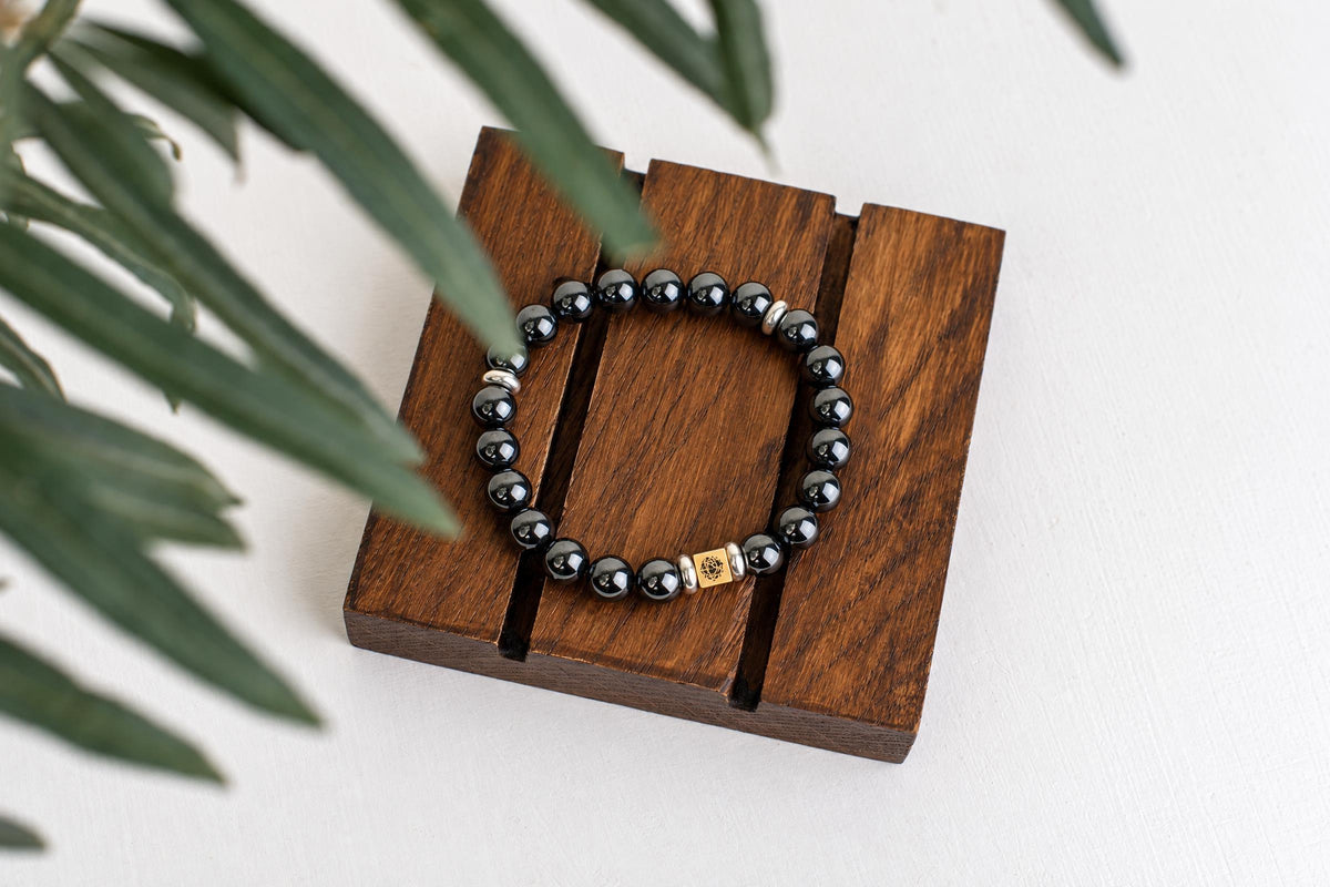 A bracelet with black spherical beads, silver-toned spacers, and a gold-coloured square charm rests on a dark wooden surface.